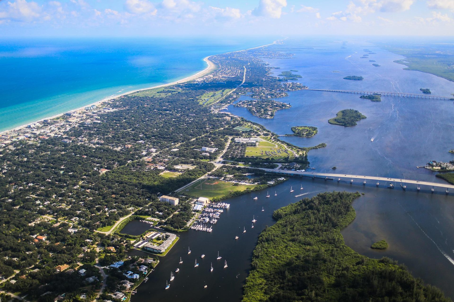 Aerial photograph of the Vero Beach barrier island in Indian River County, Florida — showing the Atlantic Ocean and white-sand beach, tree-canopied Central Beach neighborhoods, the Indian River Lagoon with sailboats near the municipal marina, and the Merrill P. Barber Bridge.