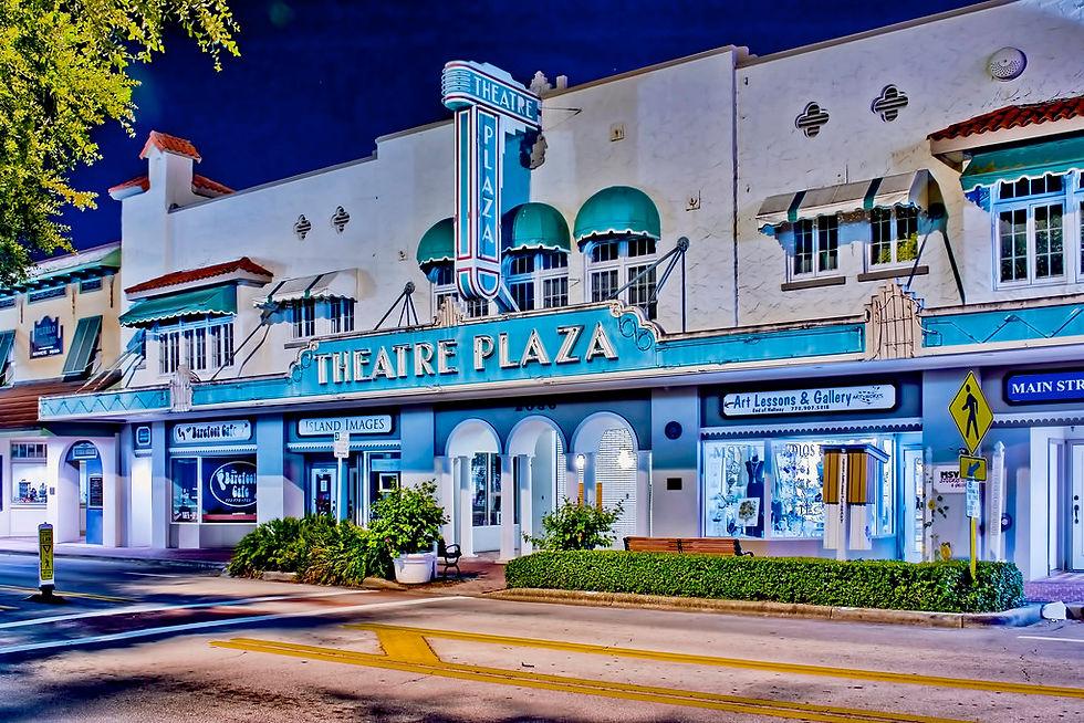 Nighttime photograph of the historic Theatre Plaza on 14th Avenue in downtown Vero Beach, Florida — a 1920s Mediterranean Revival building with a lit vertical THEATRE marquee, turquoise awnings, Barefoot Café, Island Images, and Art Lessons & Gallery storefronts along Main Street.