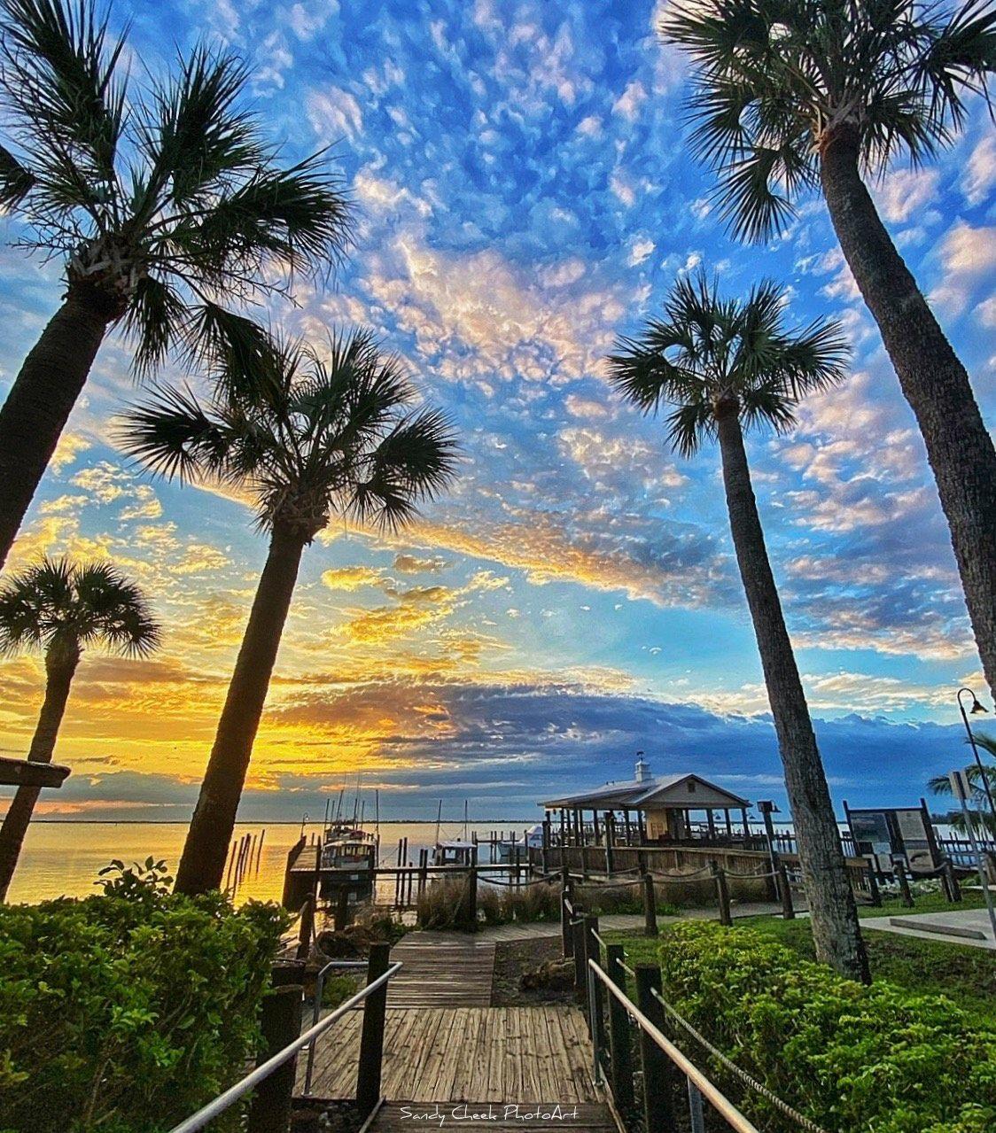 Sunset photograph at the Sebastian, Florida riverfront — a wooden boardwalk framed by tall cabbage palm trees leads to a covered gazebo and working docks on the Indian River Lagoon, with a fishing boat, sailboats, and pilings silhouetted against a gold and blue sky.